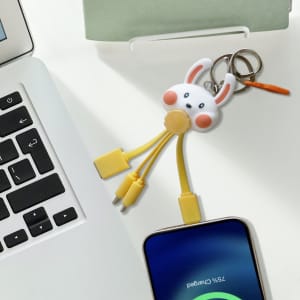 A custom-shaped PVC charging cable in yellow with a bunny keyring, resting on a desk next to a laptop and smartphone.