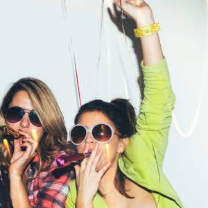 Two women wearing sunglasses, celebrating with party blowers and a yellow stretchy wristband on one wrist.