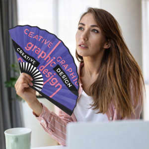 A bamboo fabric hand fan in purple and black, featuring graphic design elements, held by a woman near a laptop.