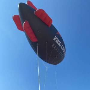 An inflatable blimp in black with red fins, floating against a clear blue sky. It features a logo.