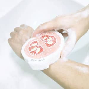 A sponge resembling a grapefruit in colour, being used with soapy hands in a sink. It has a circular shape.