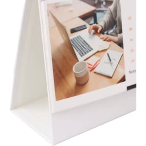 A desk calendar in a white stand, featuring a photo of a workspace with a laptop, notebook, and cup.