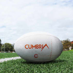 A white rugby ball with an orange logo on a grass field under a cloudy sky.