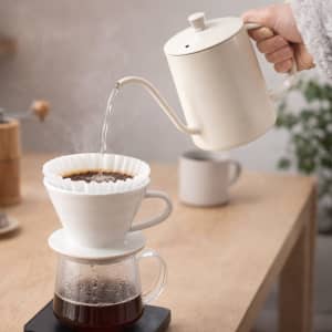 A stainless steel pour over kettle in cream colour, with a slim spout, pouring hot water over coffee in a dripper.
