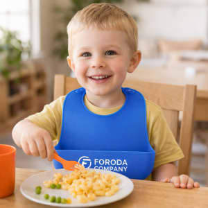 A blue silicone bib with snap closure worn by a smiling child, eating pasta and peas at a dining table.