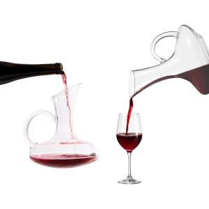 A glass handled wine decanter is pouring red wine into a glass, with a wine bottle positioned nearby.
