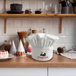 A white chef hat with a logo on a kitchen countertop, surrounded by kitchenware and decorations.