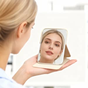 A portable mini mirror in a light gold casing, held by a person, reflecting their face.