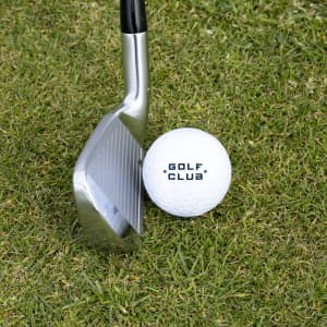 A golf club rests near a white golf ball on green grass, featuring a logo on the ball.