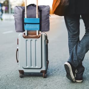 A grey travel compressed bag attached to a silver suitcase, being wheeled along a pavement by a person in dark clothing.