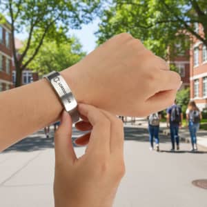 A metal bracelet being adjusted on a wrist, set against a background of trees and a campus pathway.