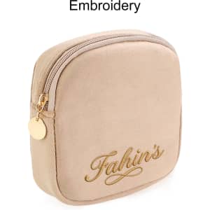 A beige mini makeup bag featuring a zip closure and gold embroidery, with a small round charm attached.