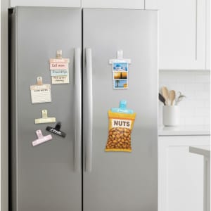 Translucent plastic magnetic clips in various pastel colours, holding notes and a snack packet on a refrigerator door.