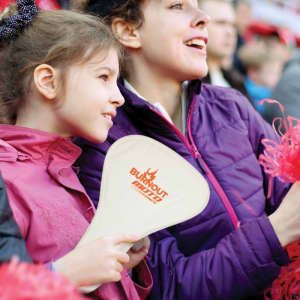 A foldable event fan in a cream colour held by a child, with a woman beside her wearing a purple jacket.
