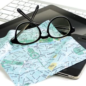 A map with a layout of Paris, black glasses, a smartphone, and a tablet on a white keyboard.