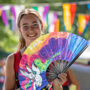 A large folding fan with a colourful design, featuring shades of blue, pink, and yellow, held by a young woman.