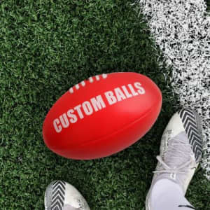 A red size 5 football with a white logo, resting on green artificial grass beside a shoe.