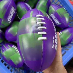 A purple and green Mini Footy Ball with white laces and a logo, held in a hand among others in a basket.