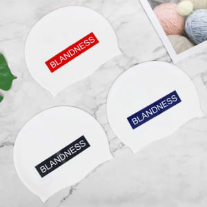 Three silicone swim caps in white, featuring red and navy text banners, displayed on a marble surface with yarn nearby.