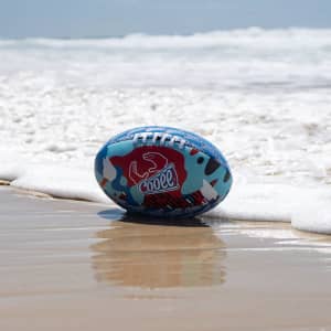 A medium soft footy ball resting on a sandy beach, featuring a blue and red camouflage design.