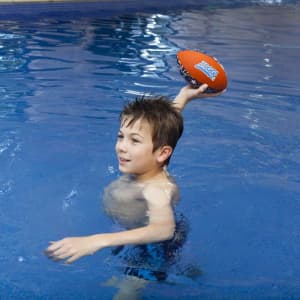 A medium soft rugby ball in orange held by a child in a pool of water, with a logo visible on the ball.