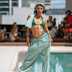 A model poses in a green bikini and mint-coloured pants, accessorised with a headscarf, near a poolside setting.