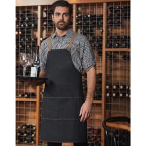 A denim bib apron in dark blue, featuring pockets and a logo, worn by a model in a wine cellar setting.