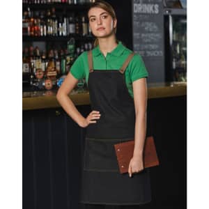 A denim bib apron in dark denim with tan straps, worn by a model holding a clipboard, set against a bar background.
