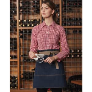 A denim half apron with a light brown belt, worn by a woman in a checked shirt, set against a wine rack background.