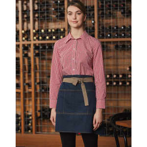 A model wears a denim half apron with a beige waistband, paired with a red checkered shirt against a wine rack backdrop.