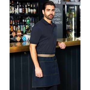 A man wearing a denim half apron stands at a bar, featuring a logo on the apron and a dark background with shelves of drinks.