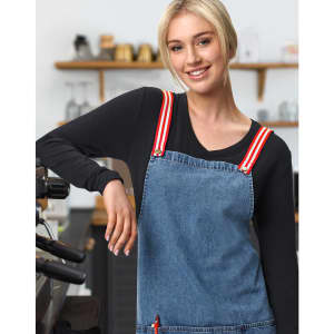 A denim bib apron with red and white striped straps, worn by a model in a café setting.