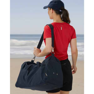 A woman wearing a red shirt and black shorts carries a navy blue duffle bag by the beach. The bag has a logo.