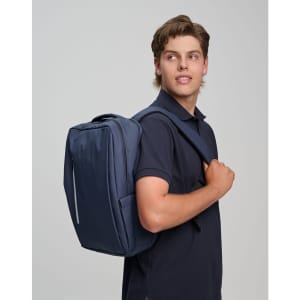 A navy blue backpack is worn over one shoulder by a young man in a navy polo shirt. The backpack features a logo.