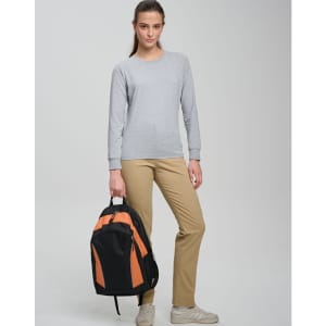 A young woman stands with an orange and black backpack, wearing a grey long-sleeve top and beige trousers.