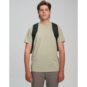 A man wearing a light khaki T-shirt and grey pants, carrying a black backpack with shoulder straps.