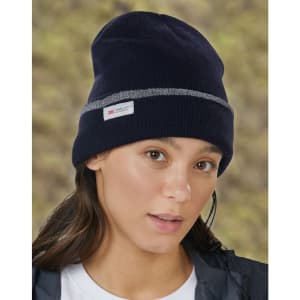 A navy beanie with a grey band and a logo, worn by a person with long hair in a blurred outdoor background.