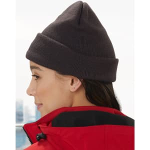 Acrylic beanie in dark grey worn by a person with long hair, paired with a red jacket. The beanie has a logo.