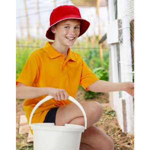 A red bucket hat sits on a boy wearing an orange polo shirt, holding a white bucket in a garden setting.