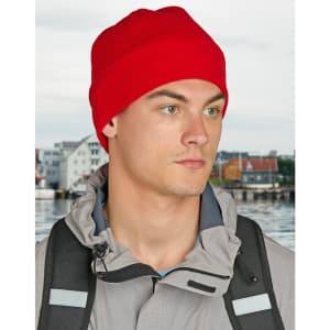 A red beanie worn by a person, paired with a grey jacket, set against a blurred background of a waterfront.