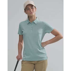 A woman wearing a light blue polo shirt and a beige cap stands with a golf club in hand. The image features a logo on the shirt.