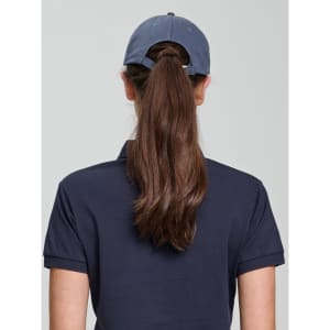 A woman with long hair wears a navy blue unstructured cap, viewed from the back. The cap features a branding logo.