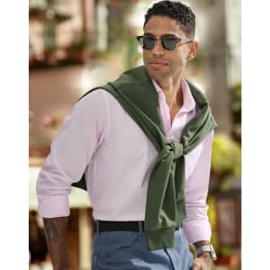 A man wearing a pink shirt and sunglasses, with a green sweater tied around his shoulders, poses outdoors.