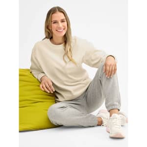 A unisex cream crew neck sweater paired with grey sweatpants, worn by a smiling model seated on a green cushion.