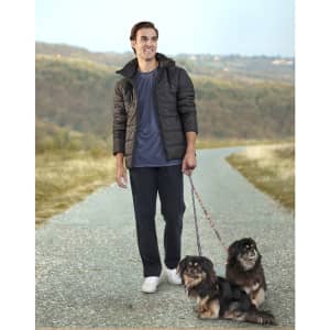 A man wearing a black sustainable parka jacket walks two dogs along a rural path, set against a hilly background.