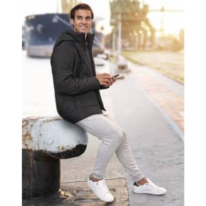 A man wearing a black unisex sustainable parka jacket, seated by a waterfront, holding a phone.
