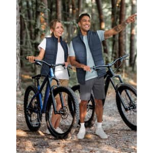 A pair of models wearing navy insulated puffer vests, standing beside bicycles in a forest setting.