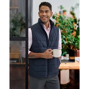 A man wearing a navy insulated puffer vest stands holding a mug, with greenery in the background.