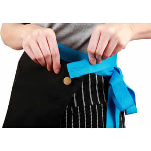 A black canvas short waist apron with a bright blue tie, being fastened around the waist.
