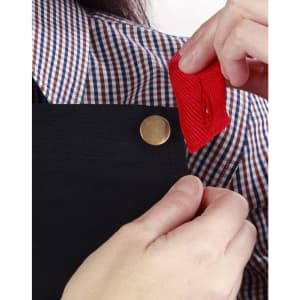 A black canvas bib apron is being adjusted, showcasing a button detail and a red fabric tag.